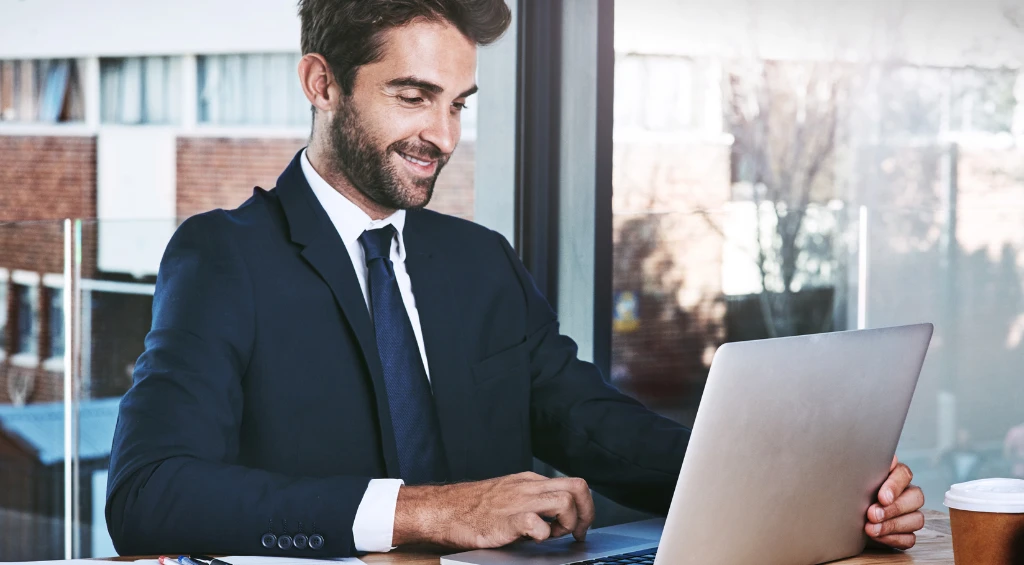 Homem de negócios sorridente em um ambiente de escritório, usando terno e trabalhando em um laptop, representando facilidade para obter crédito para MEI.