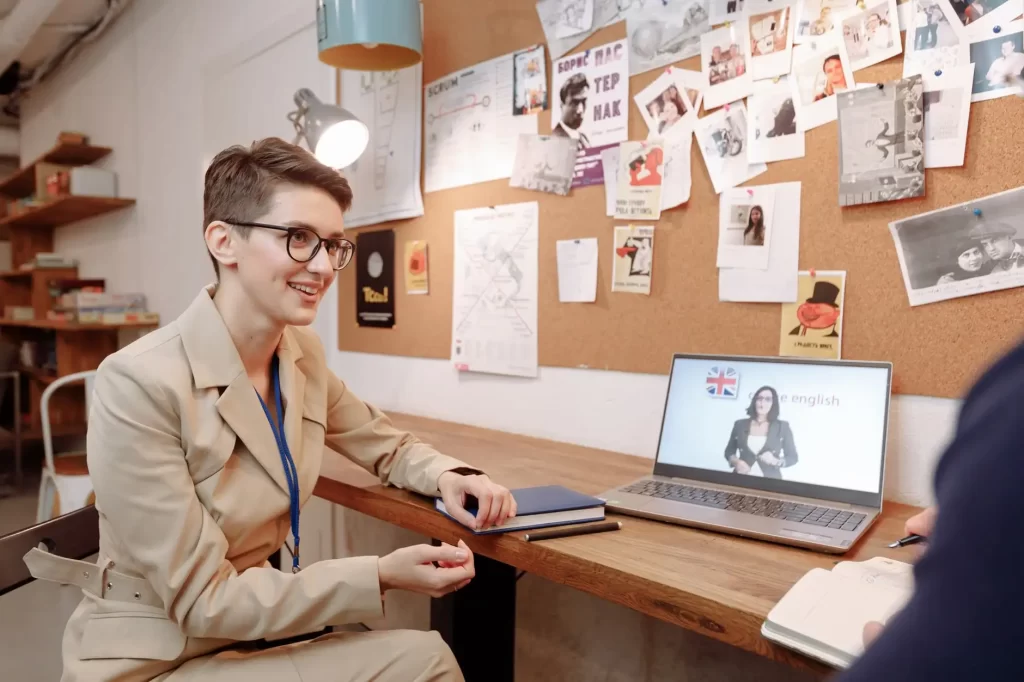Instrutor de inglês em sala de aula, com aluno conversando, usando notebook com bandeira do Reino Unido e materiais fixados no quadro de cortiça. Foto sobre cursos de inglês baratos online: aprenda sem gastar muito.
