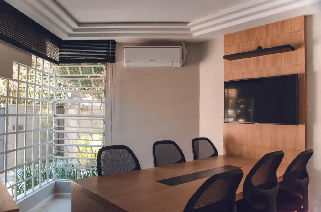 Ambiente corporativo com ar-condicionado split instalado acima de mesa de reuniões, ao lado de painel de TV, áreas com ventilação e climatização para conforto