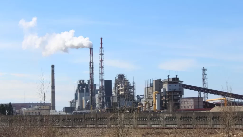 Vista de uma grande planta industrial com chaminés e estruturas metálicas ao fundo, em ambiente externo, com céu azul e vegetação ao primeiro plano, representando segurança e adequação NR-12 em Curitiba e região na campanha Sua Indústria Segura 2026.